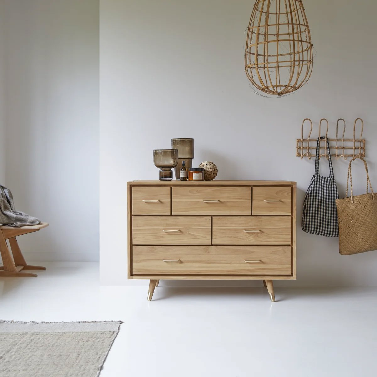Cómoda de poca altura en madera clara y cajones amplios con dos piezas de cristal como adorno sobre ella. Además, se ve una lámpara colgante en fibras naturales y una percha también en fibras naturales en la pared. 
