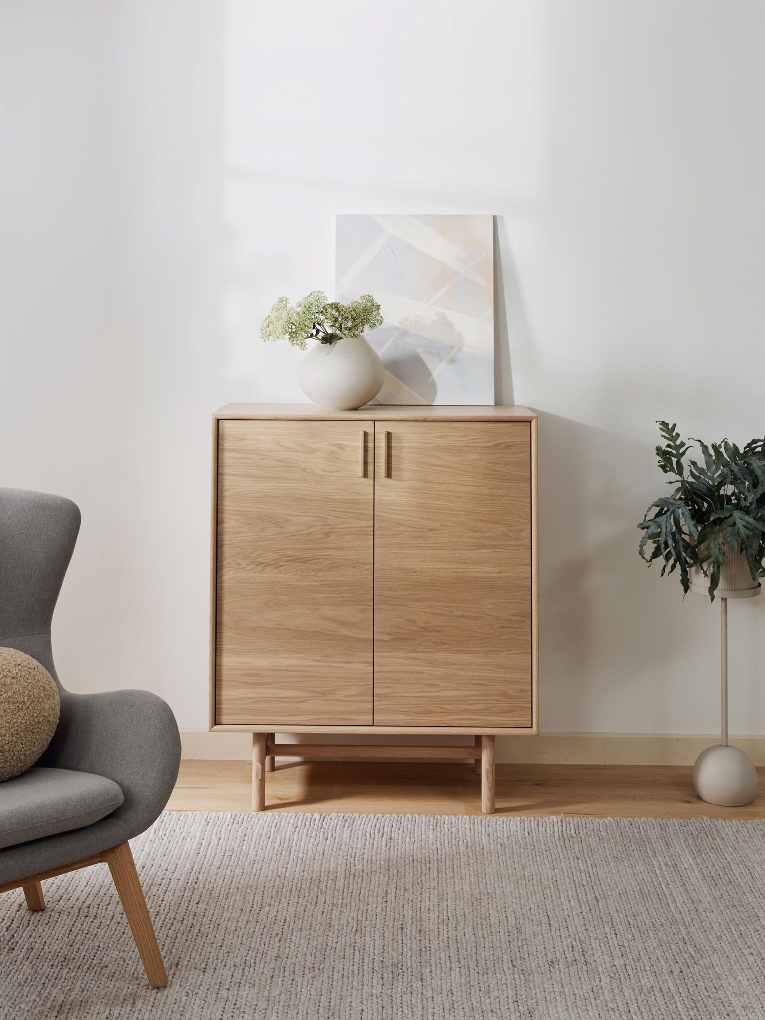 Cómoda de dos puertas color madera claro con jarrón blanco sobre ella con planta verde pequeña. También se ha puesto en la superficie un cuadro abstracto de colores pasteles muy claros. 
